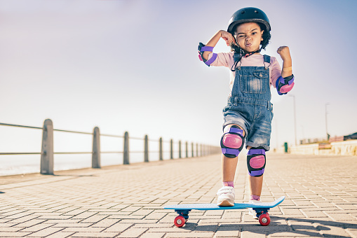 Children, skate and strong with a girl on her skateboard for fun on the beach promenade alone during summer. Muscle, bicep and helmet with a female child skating outside on a sidewalk with knee pads