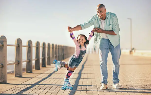 Photo of Skating, learning and father holding hands of his child while teaching her on the beach promenade. Family, love and dad helping his young girl kid for support, balance and care to outdoor rollerskate