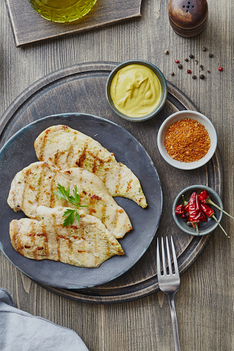 Traditional chicken breast barbecue on a plate, with mustard, black pepper, chili pepper and olive oil, served on a dark rustic wooden bar, restaurant or home kitchen table, top view with copy space