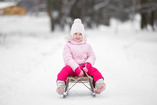Happy smiling little girl in pink warm clothes sitting on wooden sledge on white snow at nature park. Child enjoying cold winter day. Front view.