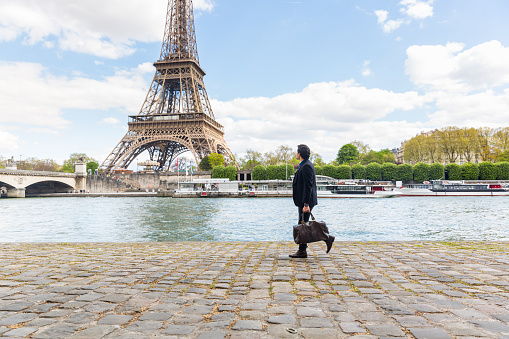 Businessman in Paris walking along the Seine looking at Eiffel tower on background - Multiracial young man carrying a bag and commuting to work during the day