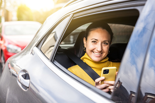 Happy woman portrait sitting on the back seat of a taxi car in the city looking out of the window - Smiling young caucasian woman using phone and car service - Travel and commuting concepts