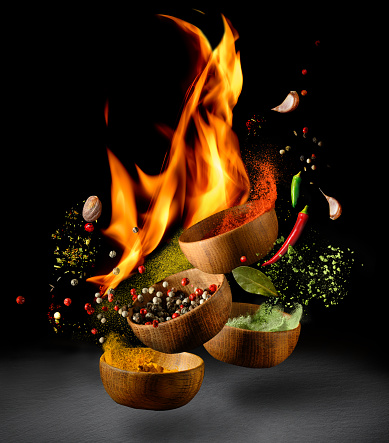 Hot spices and chili peppers in wooden bowls flying over black background and fire. Spices and seasonings powder with flame splash. Freeze motion photo.