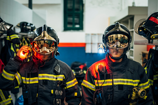 A team of three multiracial firefighters standing in protective suits and gear in front of two fire rescue trucks.  They are a diverse group, including a woman and an African American man, smiling at the camera.