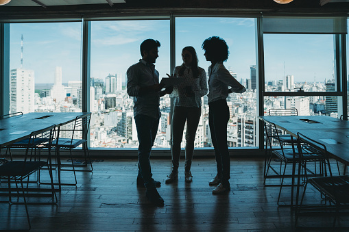 Silhouette of three business people in a modern office. Skyline of the city in the background. They are brainstorming together.