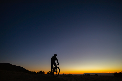 a man in silhouette mountain bikes underneath a clear twilight sky.  such wide open beautiful nature scenery and outdoor activity can be found in the sandia mountains of albuquerque, new mexico.  horizontal wide angle low angle composition.