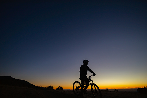 a man in silhouette mountain bikes underneath a clear twilight sky.  such wide open beautiful nature scenery and outdoor activity can be found in the sandia mountains of albuquerque, new mexico.  horizontal wide angle low angle composition.