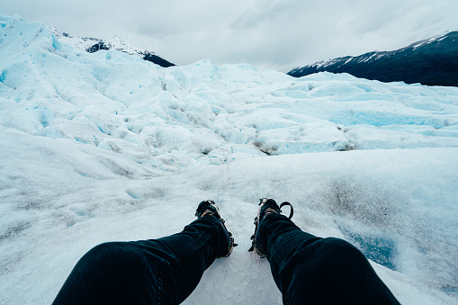 Pov view of legs and shoes with crampons on Perito Moreno glacier, Patagonia. A young adult man is resting during a hike on the glacier.