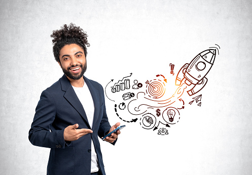 Middle eastern businessman happy portrait pointing at smartphone in hand. Black doodle sketch with start up plan on grey concrete wall, graphs and rocket launch. Concept of project