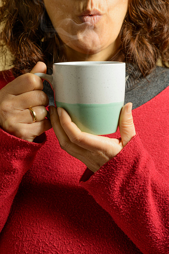 A woman in a red jersey blows to cool the hot beverage in the cup she holds in her hands.