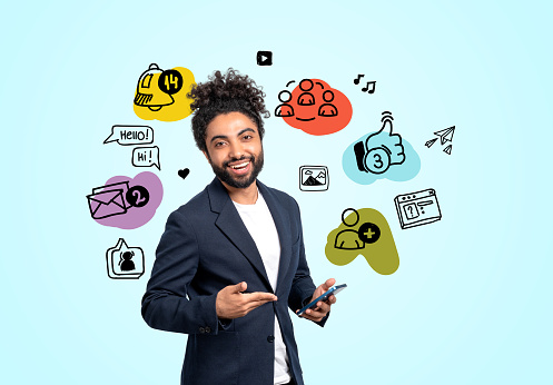 Smiling arab businessman hand pointing at smartphone, colorful social media doodle icons on blue background. Concept of online communication and marketing