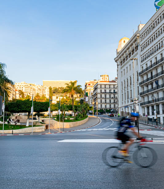 Motion blur bicycle rider in Zighoud Youcef Boulevard. Algiers, Algeria - October 28, 2022:  Central post office place and building, Algerian flag posts, palm trees and the El Aurassi Hotel in background with golden sunlight  in the morning. algeria flag silhouettes stock pictures, royalty-free photos & images