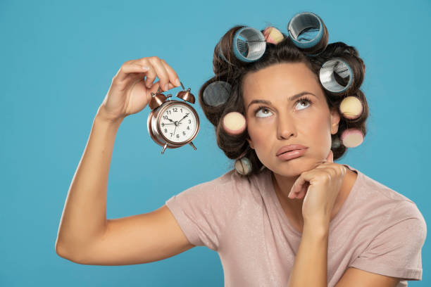 beautiful bored woman with hair curlers holding a clock on a blue background - waiting women clock boredom imagens e fotografias de stock