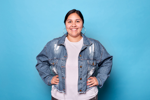 Young curvy latina woman wearing denim jacket and hoop earrings, smiling looking at camera isolated on blue background. Copy space.
