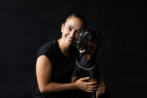 Front view of caucasian female with toothy smile looking at camera and hugging Doberman pinscher dog  on black background.