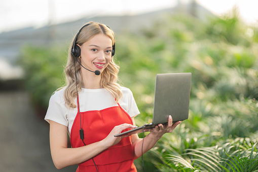 Young woman with call center headset is hanging a laptop. She is looking at camera. She is standing among green plants.