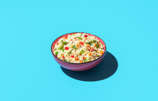Bowl with fried rice and vegetables minimalist on a colorful table. White rice with pieces of carrots, red pepper and green beans in a red bowl.