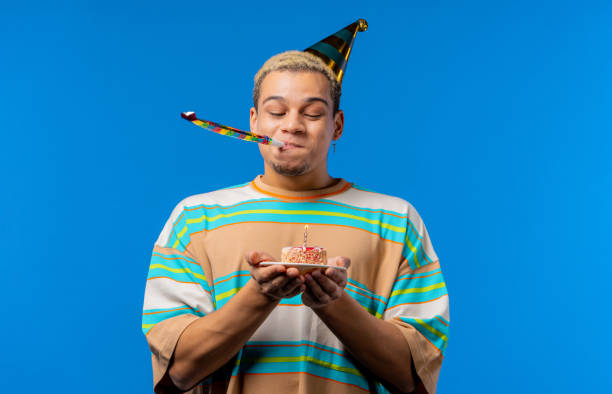 istock Happy birthday man making wish - blowing candle on cake. Curly haired guy laughs, celebrating anniversary. Young teenager on blue background. 1456884925