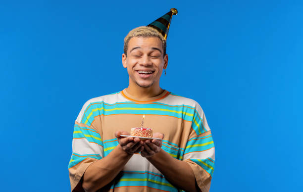istock Happy birthday man making wish - blowing candle on cake. Curly haired guy laughs, celebrating anniversary. Young teenager on blue background. 1456885296
