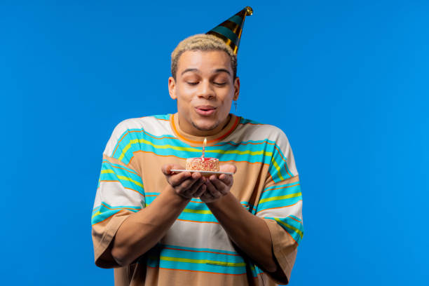 istock Happy birthday man making wish - blowing candle on cake. Curly haired guy laughs, celebrating anniversary. Young teenager on blue background. 1456886427