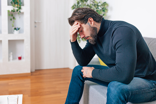 A young Caucasian man is sitting on a sofa with his head in his hand, experiencing depression.