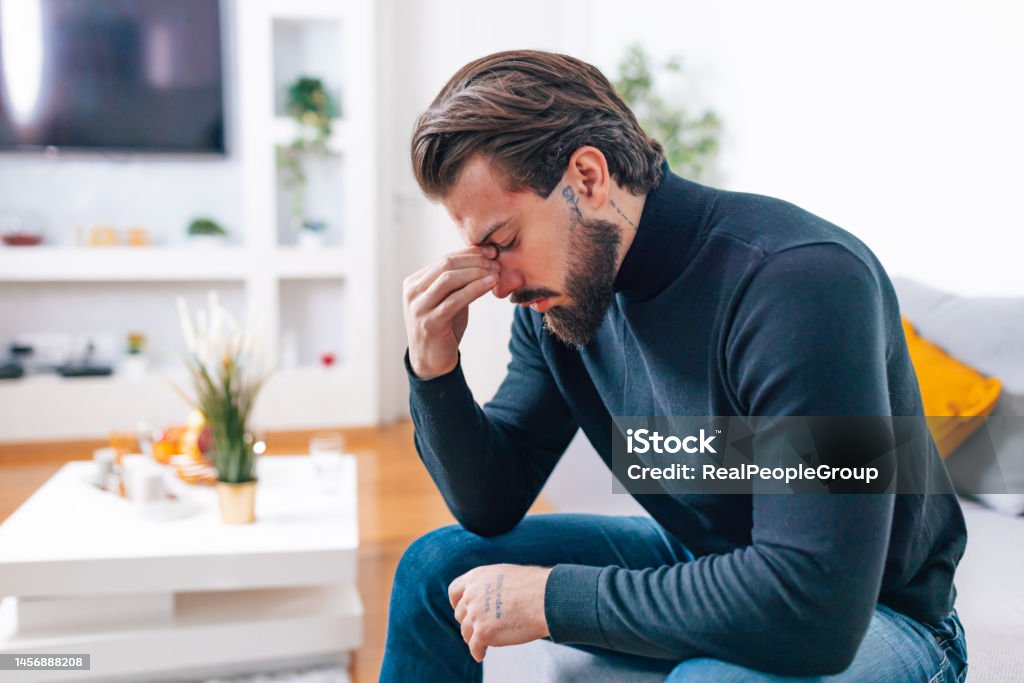 My sinuses are killing me A young Caucasian man is sitting on a sofa, holding his painful sinuses. Forehead Stock Photo