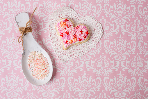 Heart-shaped cookies decorated with pink and red frosting and sprinkles.  Background is a pattered pink and white paper.