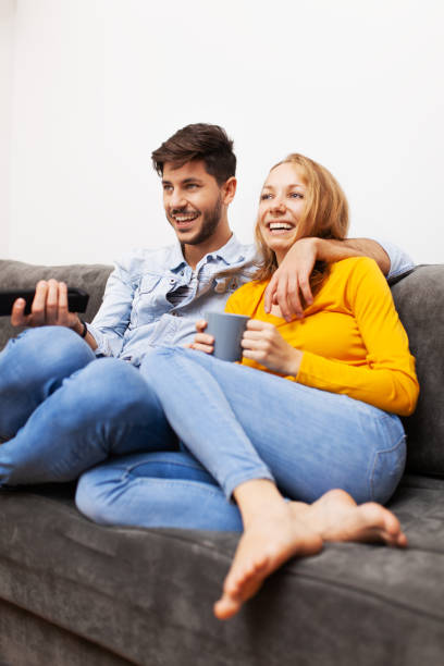couple in love on a sofa at home, watching tv and smiling stock photo