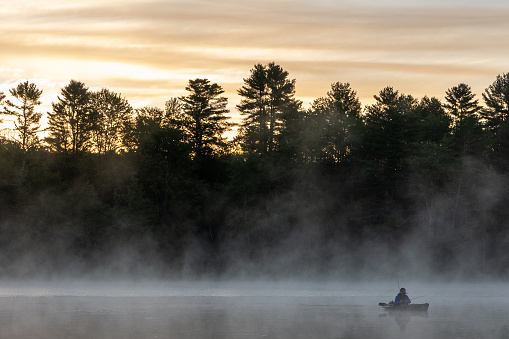Several kayakers were drifting quietly just offshore on a still lake at sunrise in northern Vermont.  The layered clouds and fog had not been burned off by the sun, and a glowing reflection of the forest is present in the calm water.