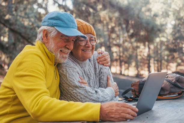 야외에서 컴퓨터 pc를 사용하는 귀여운 중년 부부의 머리 사진 초상화는 주변에 나무가 있는 자연의 숲에 있는 나무 테이블에 앉아 있습니다. - mature couple action senior couple senior adult 뉴스 사진 이미지