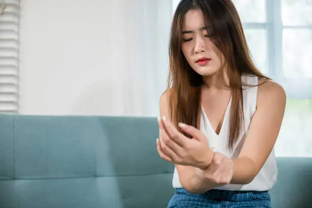 Closeup hand. Asian young woman sitting on sofa holds suffering her wrist at home, female hand injury feeling joint pain, performing self-massage, Health care and medical concept