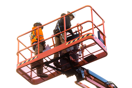 Two Caucasian construction workers operate a heavy duty aerial lift on a job site.  They follow safety protocol, wearing protective harnesses and hardhats.