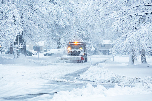 A town highway department snowplow dump truck is plowing snow and spreading road salt to clear a suburban residential district street during an extreme weather winter blizzard snow storm.