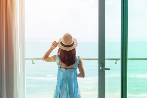 Happy woman wearing blue dress and hat looking outside window to ocean view in morning. Relax, vacation, time to travel and freedom concept