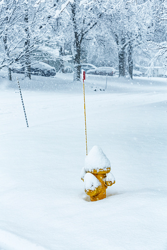 A yellow fire hydrant with a tall red locator flag is being buried under deep snow during a very heavy February blizzard storm. It is located at the edge of a corner at a road intersection between two suburban residential district streets.