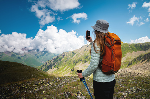 A beautiful young woman with blond hair photographs a mountain landscape on a phone camera while walking, rear view. Tourist with a backpack, vacation in the mountains.