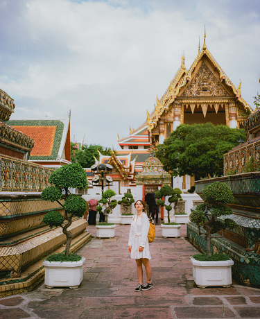 Young Caucasian woman in white dress with yellow backpack exploring Wat Pho temple in Bangkok