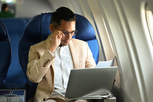 Focused asian businessman working on laptop, using wireless connection on board in an airplane during his business travel.