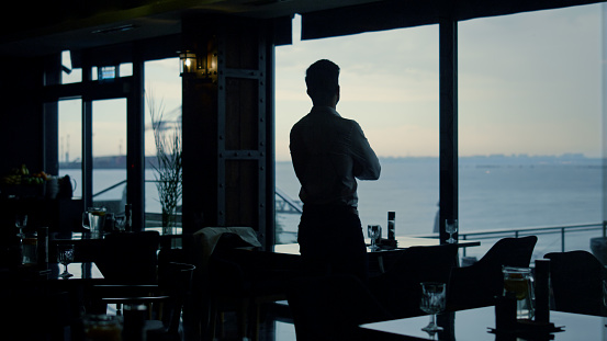 Pensive man silhouette standing restaurant hall alone. Attractive guy enjoying beautiful ocean view in panoramic lounge bar. Thoughtful boss looking window in dark interior. Business success concept.
