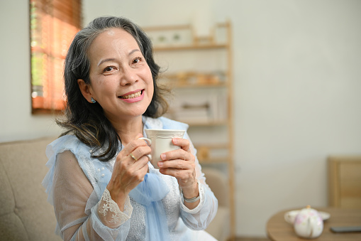 Charming and beautiful 60s aged Asian woman is in the living room holding a cup of tea, smiling and looking in the camera.