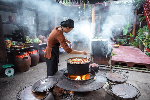 side view on young female chef cooking steamed fish in huge pod on open fire in restaurant at countryside in north vietnam