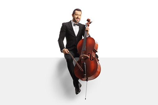 Full length portrait of a musician in a black suit and bow-tie sitting on a white panel with a cello isolated on white background