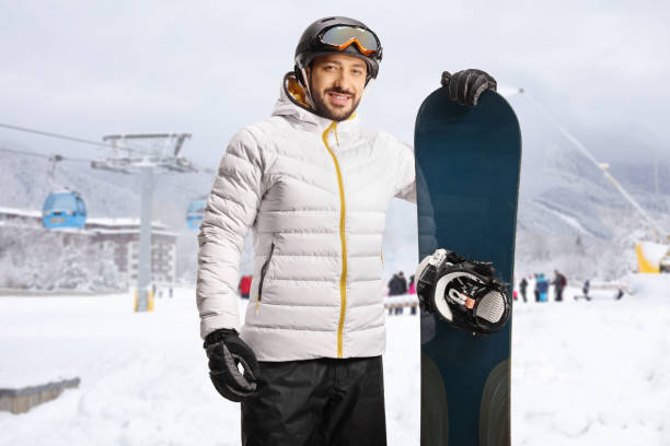 istock Smiling young man with a snowboard posing at a ski resort with a cable car 1457045515