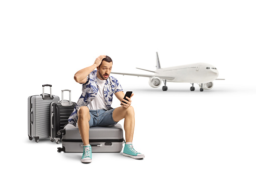 Shocked male tourist sitting on a suitcase and looking at a smartphone in front of an airplane isolated on white background