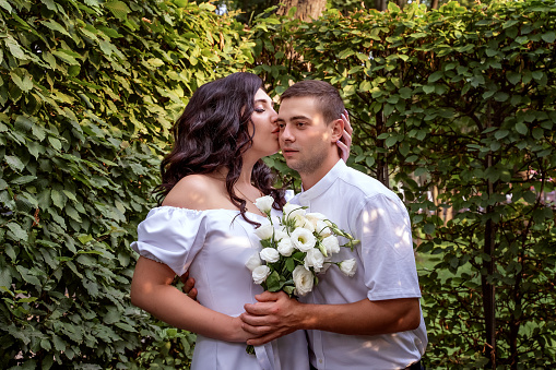 Girl in white dress gently hugs her boyfriend's neck and kisses him on the cheek. Newlyweds in the park. Portrait of beautiful couple close-up. Summer kiss of the Ukrainian beauty.
