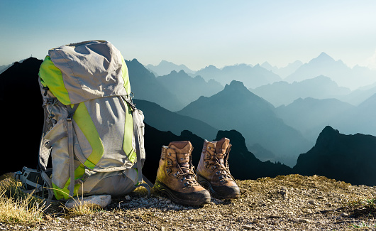 Hiking equipment with blue mountain silhouettes. Backpack and hiking shoes on top of the mountain. Beautiful view to mountain ranges and fog filled valleys. Tirol, Austria, Allgaeu, Alps.