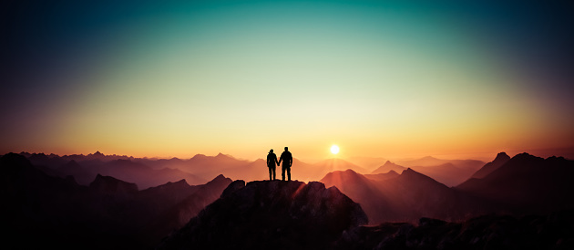 Happy winning success man and woman at sunset or sunrise standing on mountain peak holding hands and are happy for having reached mountain top summit goal during hiking travel trek. Tirol, Austria. Allgäu, Bavaria.