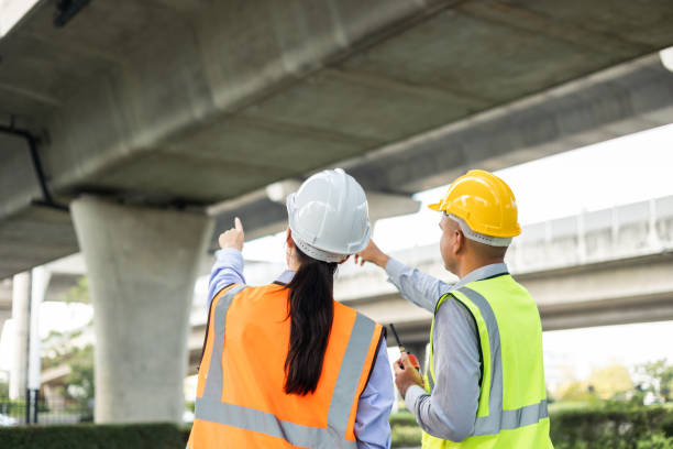 istock Two Asian worker engineer man and woman architect looking construction with white safety helmet in construction site. Standing at highway concrete road site. Work planning with blueprint and tablet. 1457049584