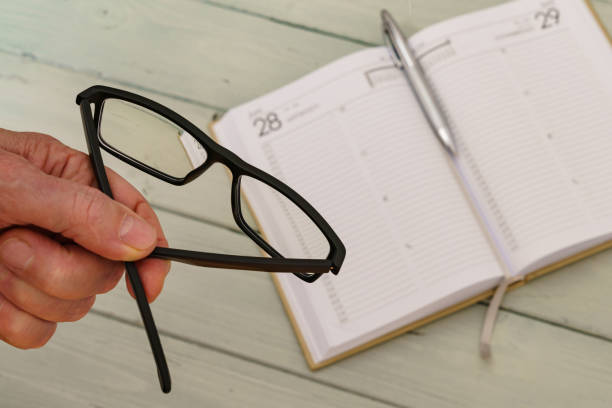 Man's hand holding glasses over a desk. On the table lies an open schedule planner out of focus.