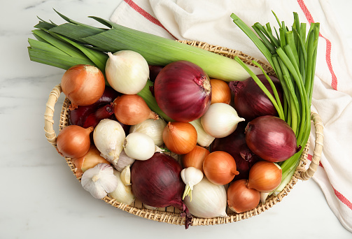 Wicker tray with fresh onion bulbs, leeks and garlic on white marble table, flat lay